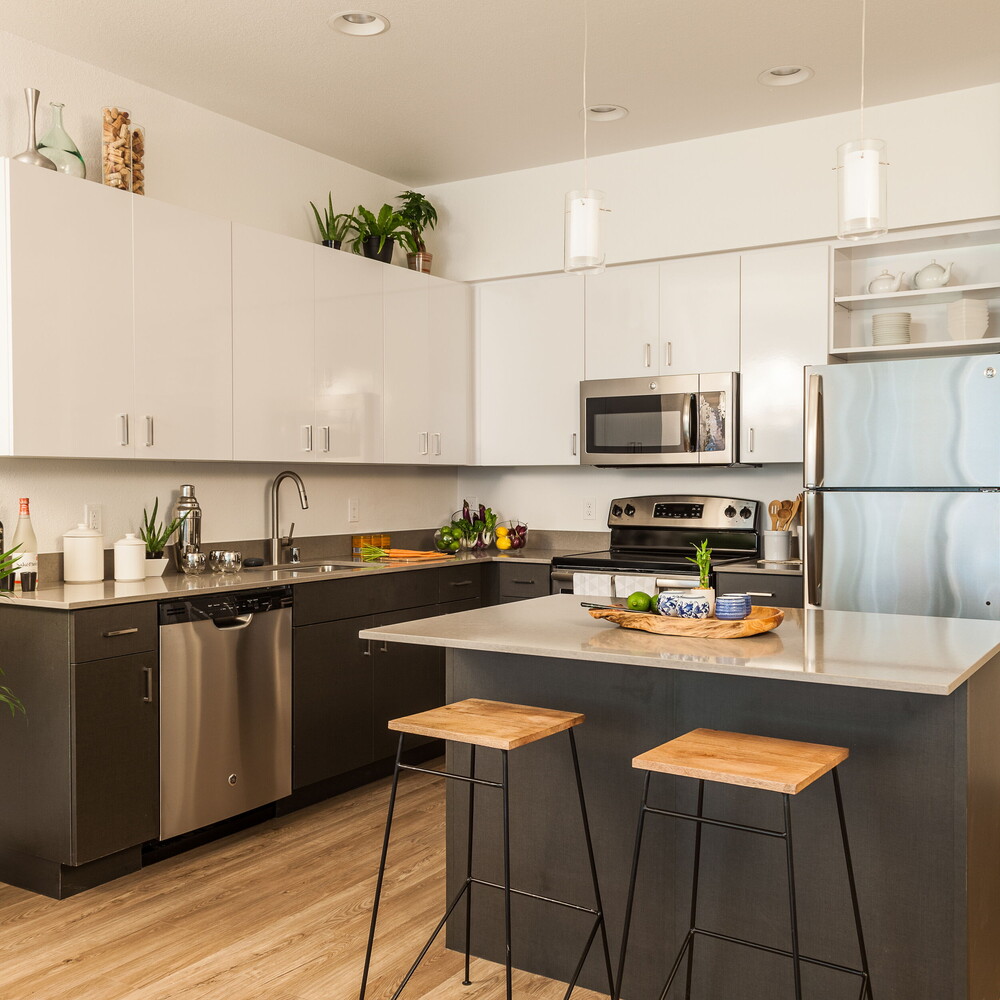 Modern fitted kitchen with island, two bar stools, stainless-steel appliances (fridge, oven, microwave), pendant lights and plants – holiday apartment.